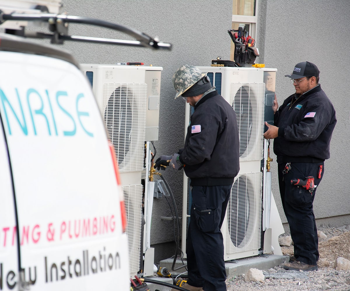 Two HVAC technicians working on heat pump units outside a building.