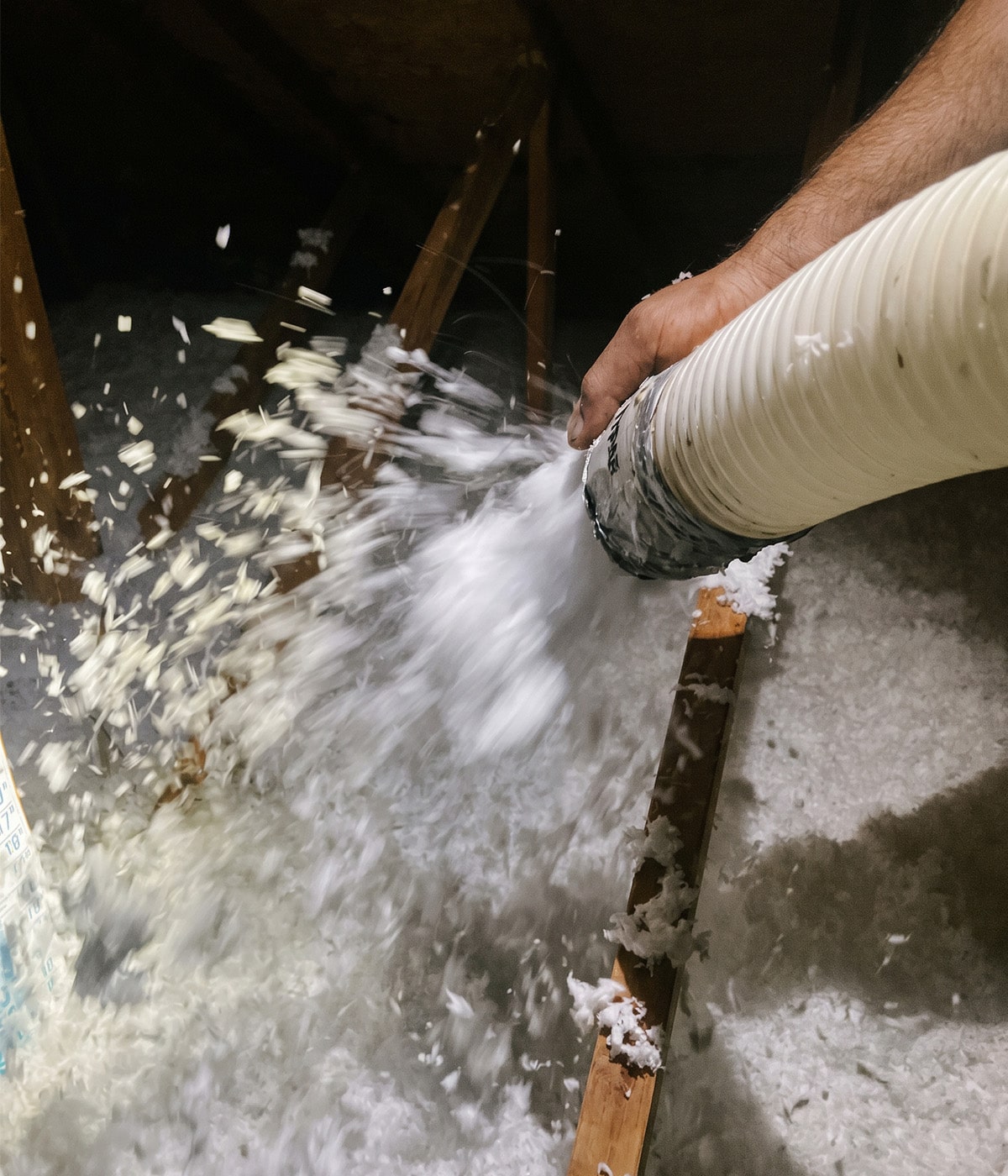 Gloveless hand guides a taped blower hose spraying white loose-fill insulation over attic joists for Sunrise Service in Las Vegas.